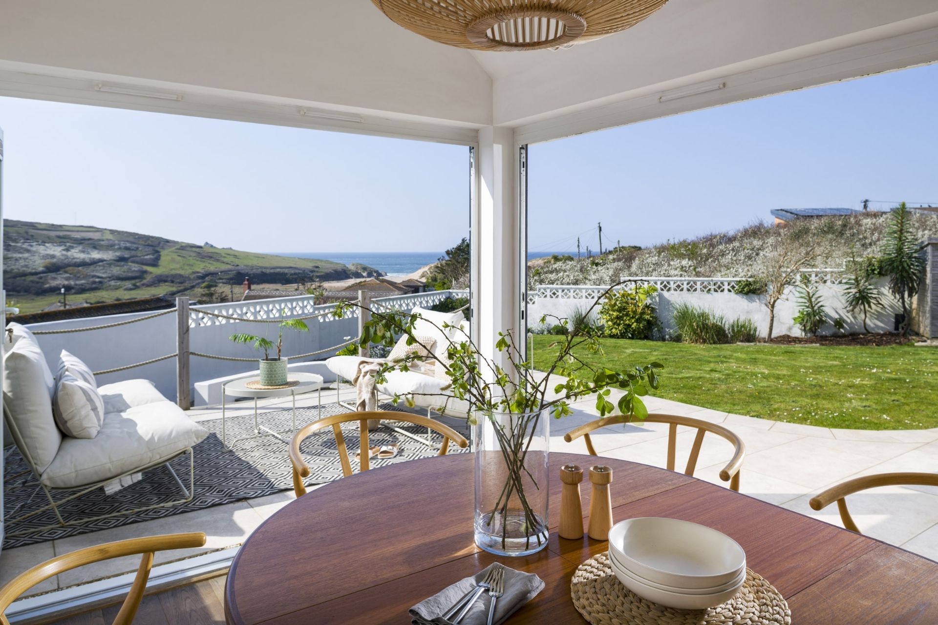 A dining area with a view of the sea, a round table, chairs, and a vase with branches.