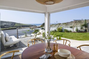 A dining area with a view of the sea, a round table, chairs, and a vase with branches. - Sunset Sands - sleeps 8 guests  in 4 bedrooms (Newquay)