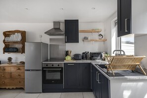 A modern kitchen with a built-in oven, a stove, and a microwave. There is a wooden shelf above the stove and a wooden chair in the corner. - Seamist  - sleeps 2 guests  in 1 bedroom (Hayle)