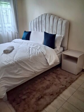 Ein ordentlich bezogenes Bett mit weißer Bettwäsche und einem aufgerollten Handtuch, ein Nachttisch und ein flauschiger Teppich auf Fliesen.