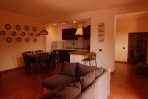 A living room with a sofa, dining table, chairs, and a kitchen area with cabinets and a stove. - Vrbo Property (Carruba-San Leonardello)