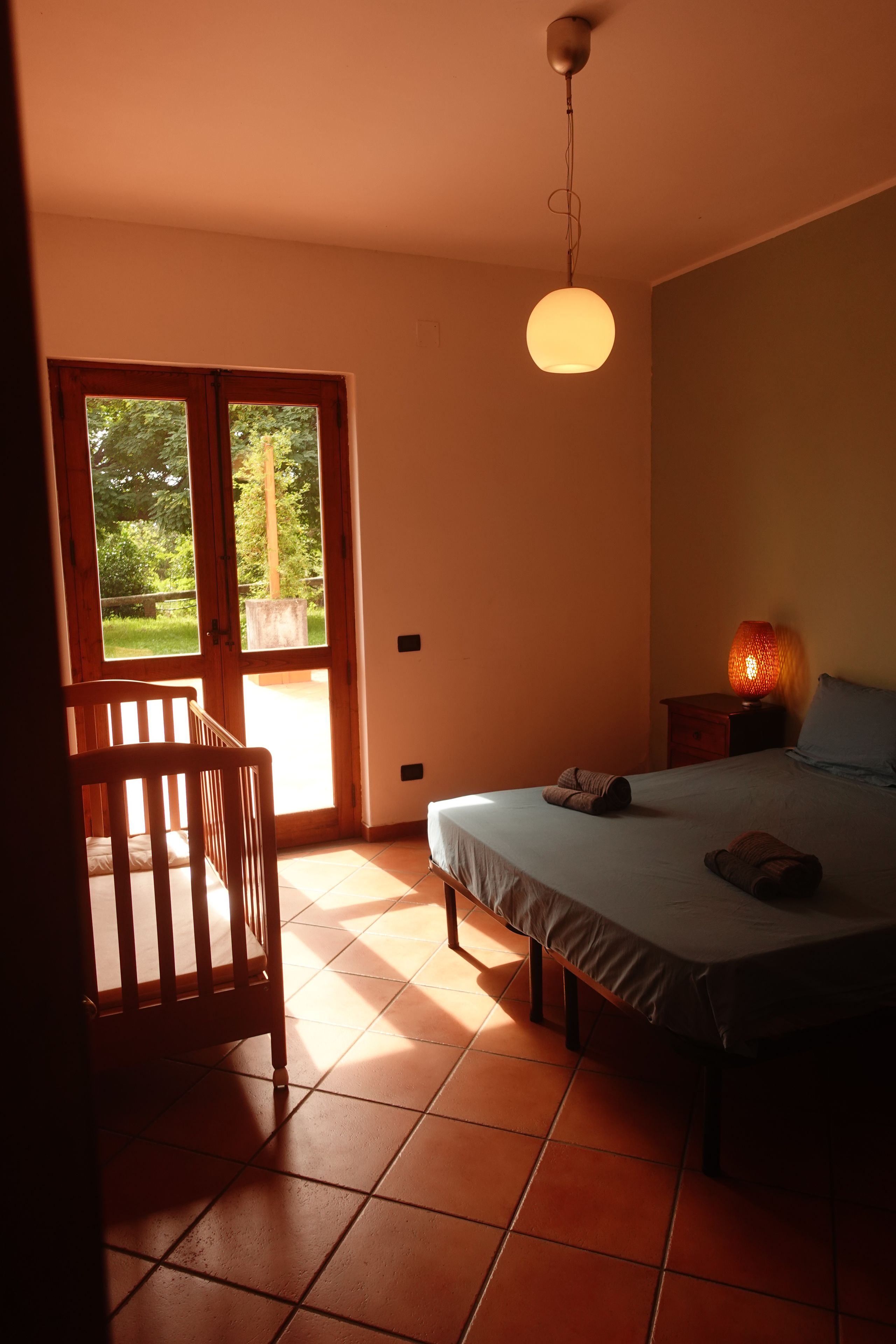 A bedroom with a bed, a chair, a bedside table with a lamp, and a window with a view of greenery.