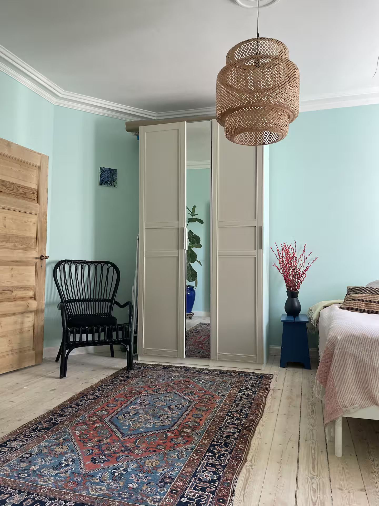 A room with a traditional design, featuring a wooden door, a black chair, a patterned rug, a bed with a pink blanket, and a hanging rattan light fixture.