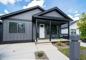 A modern two-story house with a grey exterior, a front porch with seating, and a visible mailbox. - 2BR | Hot Tub + Ecusta Trail | Free Tix (Hendersonville)