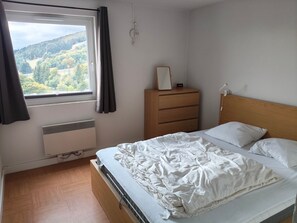 A bedroom with a bed, a dresser, a window with curtains, and a view of a forested area. - Charming bright apartment - 8 guests (Besse-et-Saint-Anastaise-Super Besse)