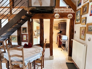 A rustic interior with exposed wooden beams, a dining area with a table and chairs, a kitchen with a stove and cabinets, and a staircase leading to an upper level. - Cosy cottage, WIFI, pets welcome - 3 guests (BAUZY)