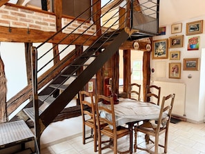 Een landelijk interieur met een houten trap, een eettafel met stoelen, en een wand versierd met kunstwerken.