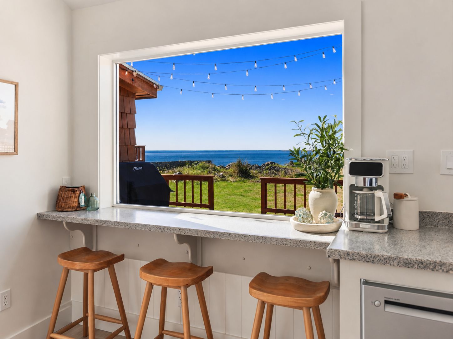 A kitchen with a large window overlooking a deck and ocean view, equipped with bar stools and a coffee maker.