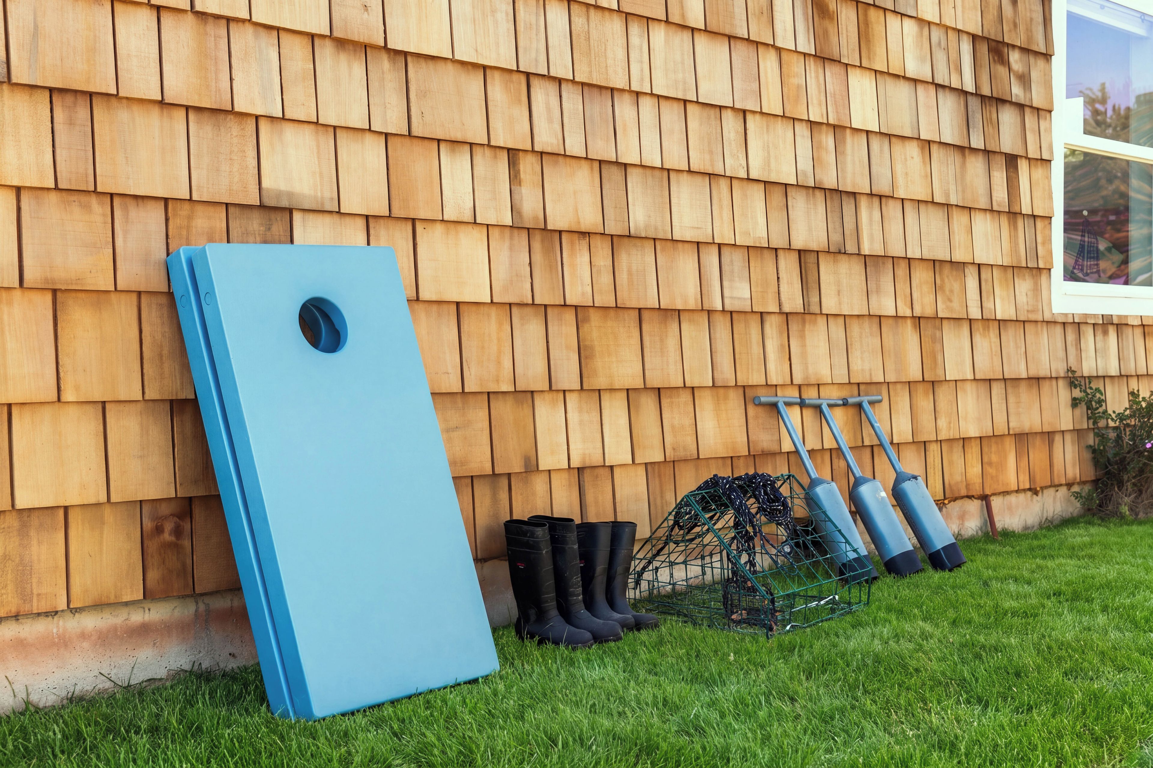 A house with a shingled exterior, a blue object leaning against the wall, and gardening tools on the grass.