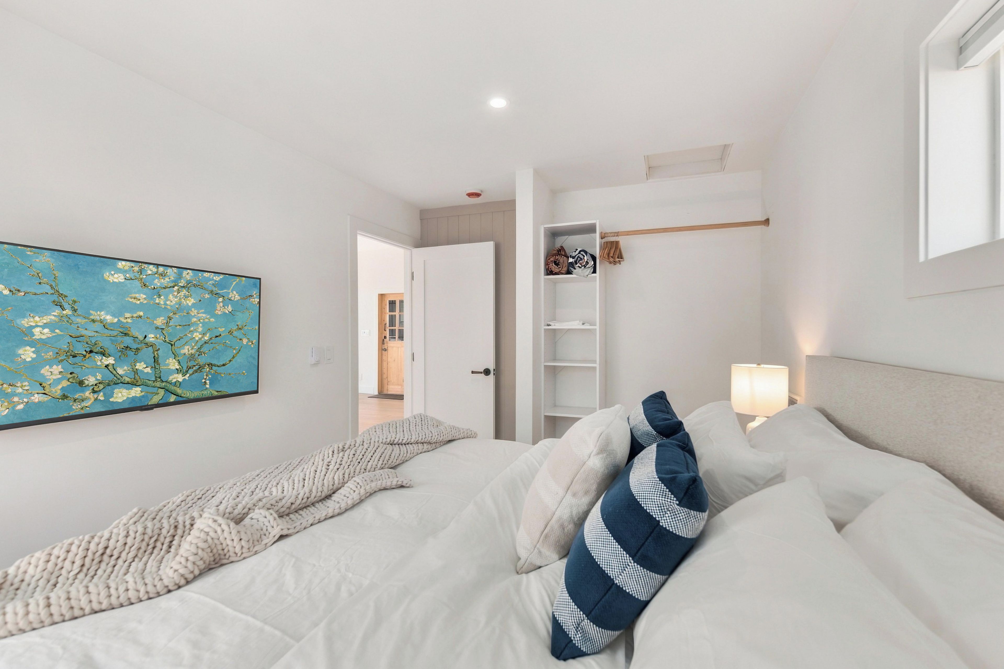 A bedroom with a bed, pillows, a painting, a wardrobe, and a nightstand with a lamp.