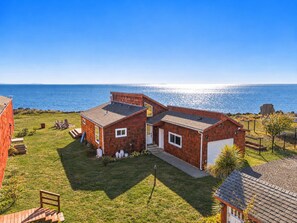 A two-story wooden house with a garage, a lawn, and a view of the ocean. - Modern Oceanfront Retreat: Beach Access, Wraparound Deck, Fireplace + Fire Pit (Tokeland)