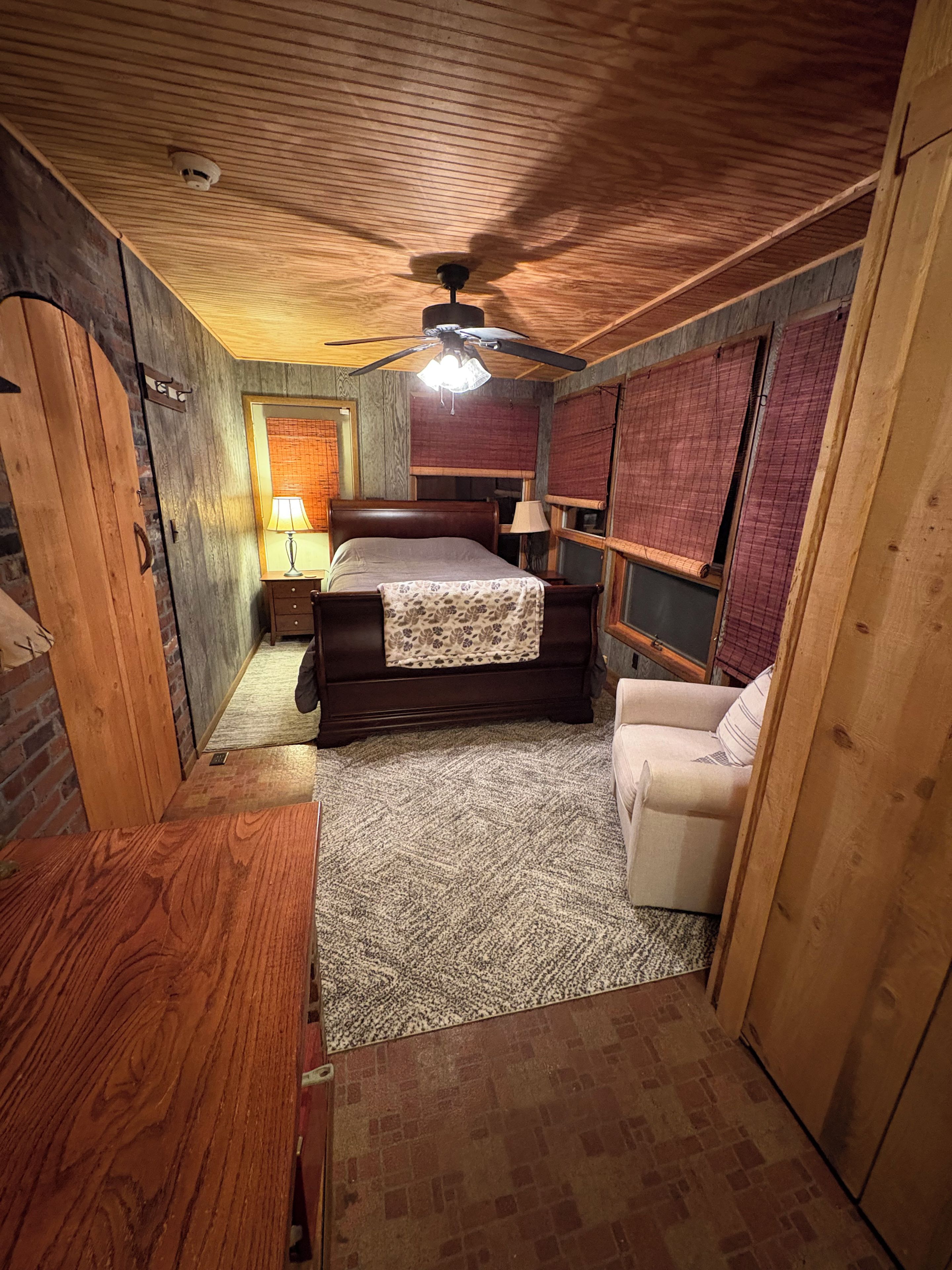 A bedroom with a wooden ceiling, a bed with a patterned bedspread, a nightstand with a lamp, a chair, and a window with blinds.