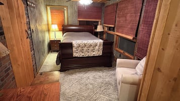 A bedroom with a wooden ceiling, a bed with a patterned bedspread, a nightstand with a lamp, a chair, and a window with blinds.