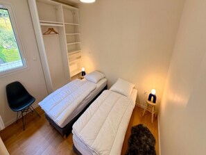 Ein kleines, gemütliches Zimmer mit zwei Einzelbetten, einem Sessel, einem Kleiderschrank und einem kleinen Tisch mit einer Lampe.