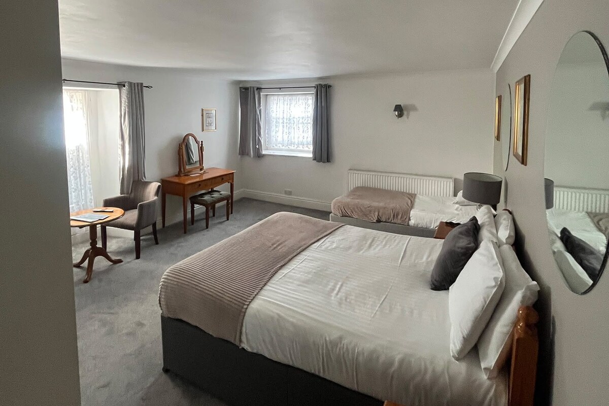 A hotel room with a large bed, a desk, a chair, a window with curtains, and a radiator.