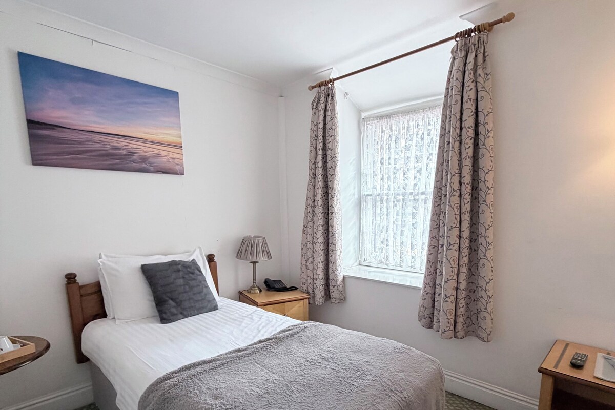 A bedroom with a bed, a nightstand, a lamp, a window with curtains, and a painting on the wall.