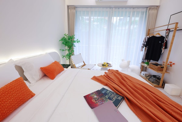 Habitación de hotel con una cama, una mesita auxiliar, una silla, una planta, un perchero y una ventana con cortinas.
