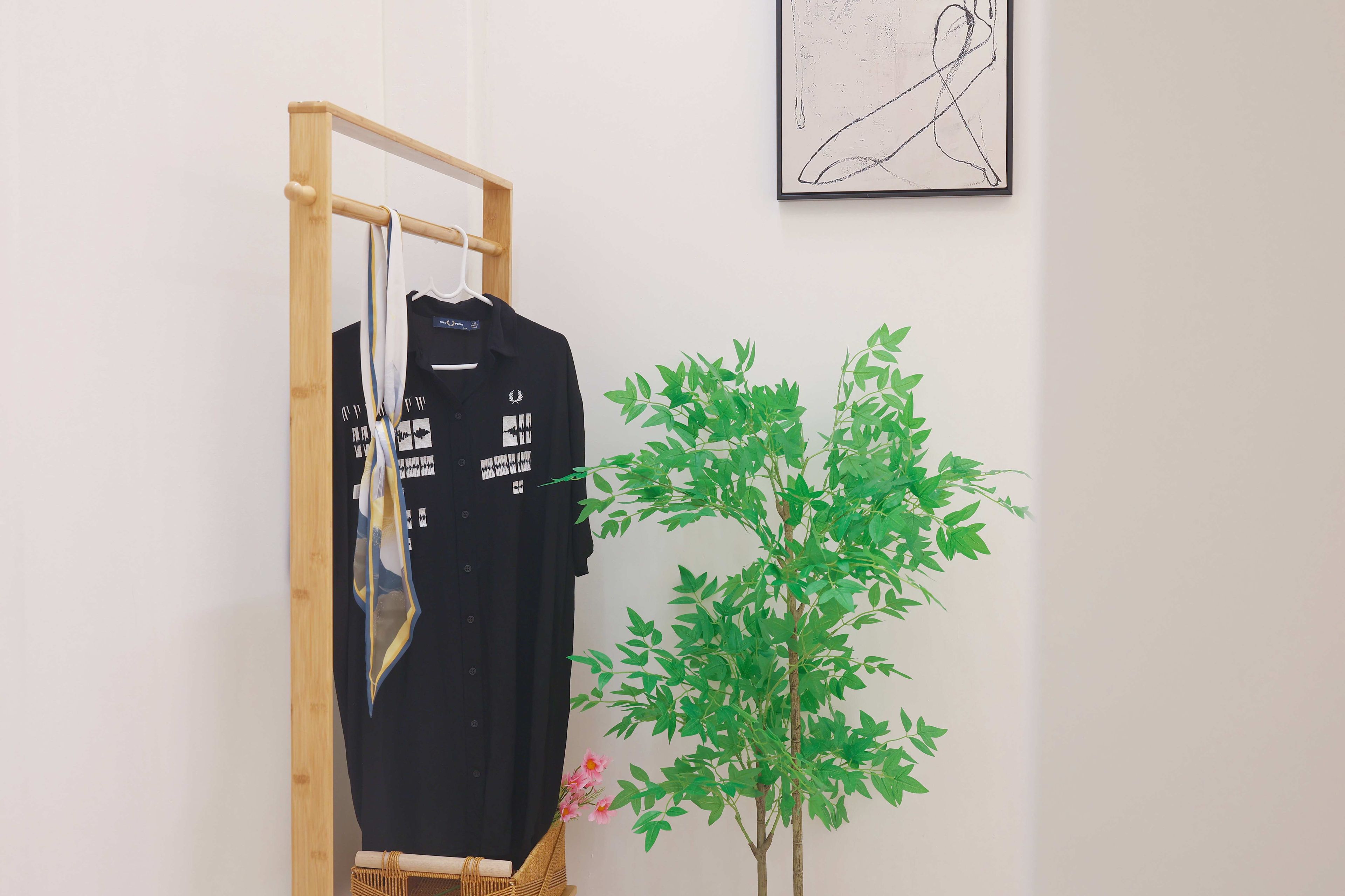 Une pièce avec un miroir à cadre en bois, un t-shirt noir orné d’un motif graphique et une plante en pot aux feuilles vertes et aux fleurs roses.