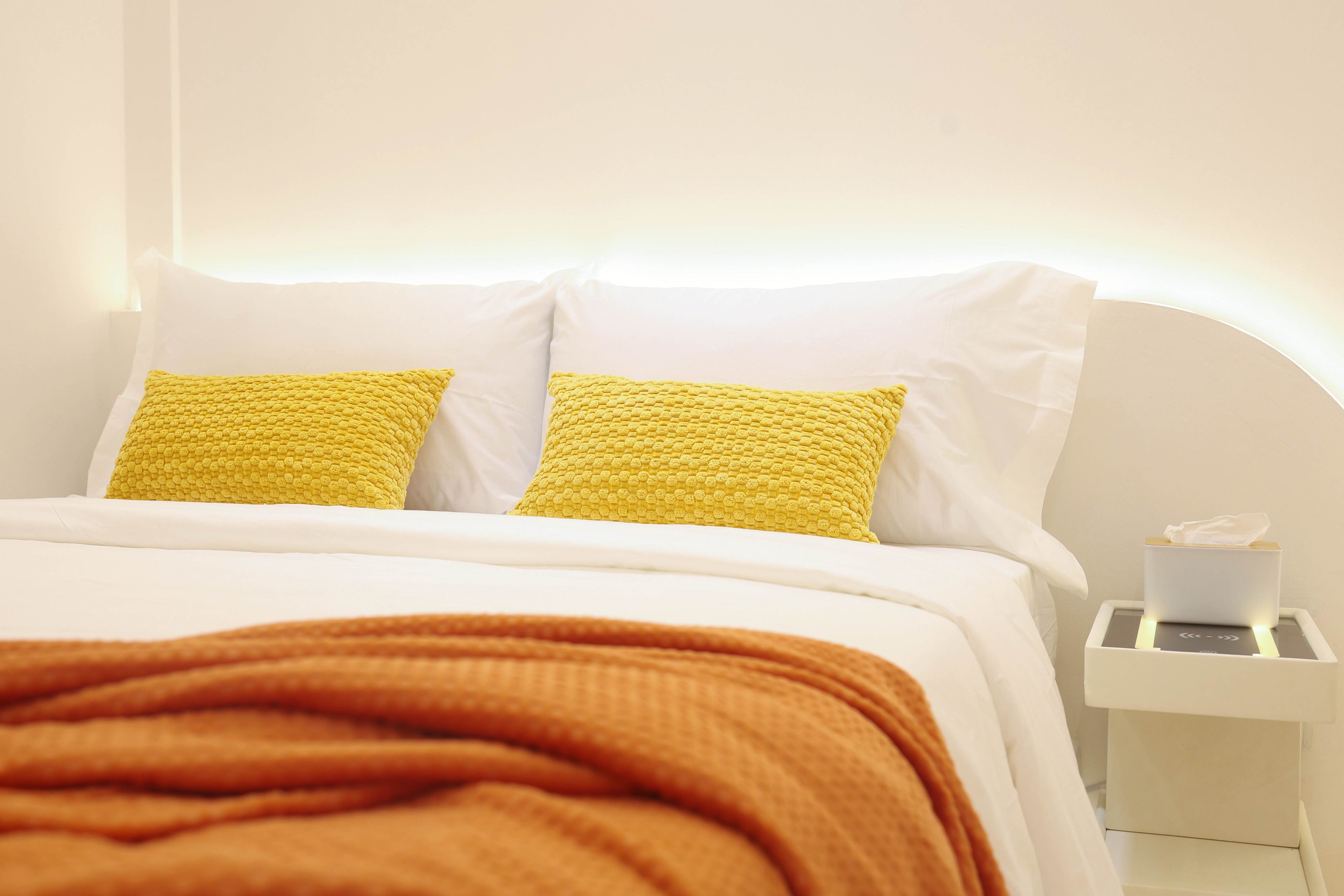 Un lit avec une literie blanche et deux oreillers jaunes, une couverture marron et une table de chevet blanche sur laquelle se trouve une boîte de mouchoirs.
