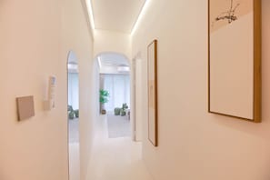 Un pasillo de diseño moderno, con piso de tonos claros, paredes blancas e iluminación empotrada en el techo. Se observan obras de arte enmarcadas en las paredes y una planta en maceta al fondo.