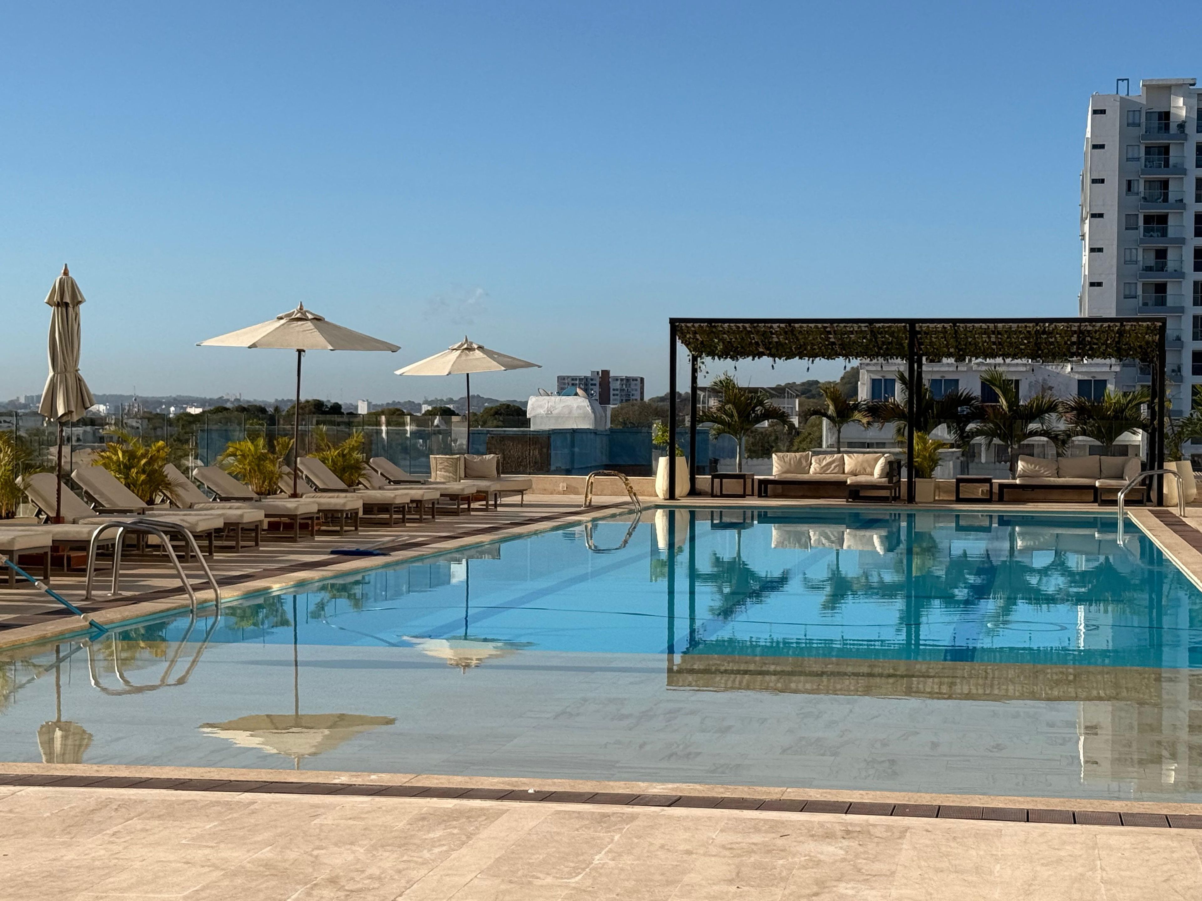 Un área de piscina en la azotea con sillones, sombrillas y una pérgola.