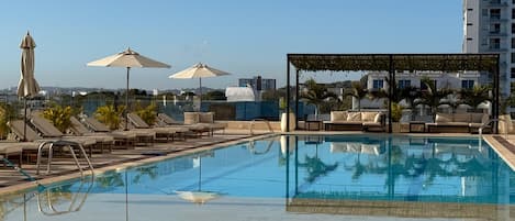 Un área de piscina en la azotea con sillones, sombrillas y una pérgola.