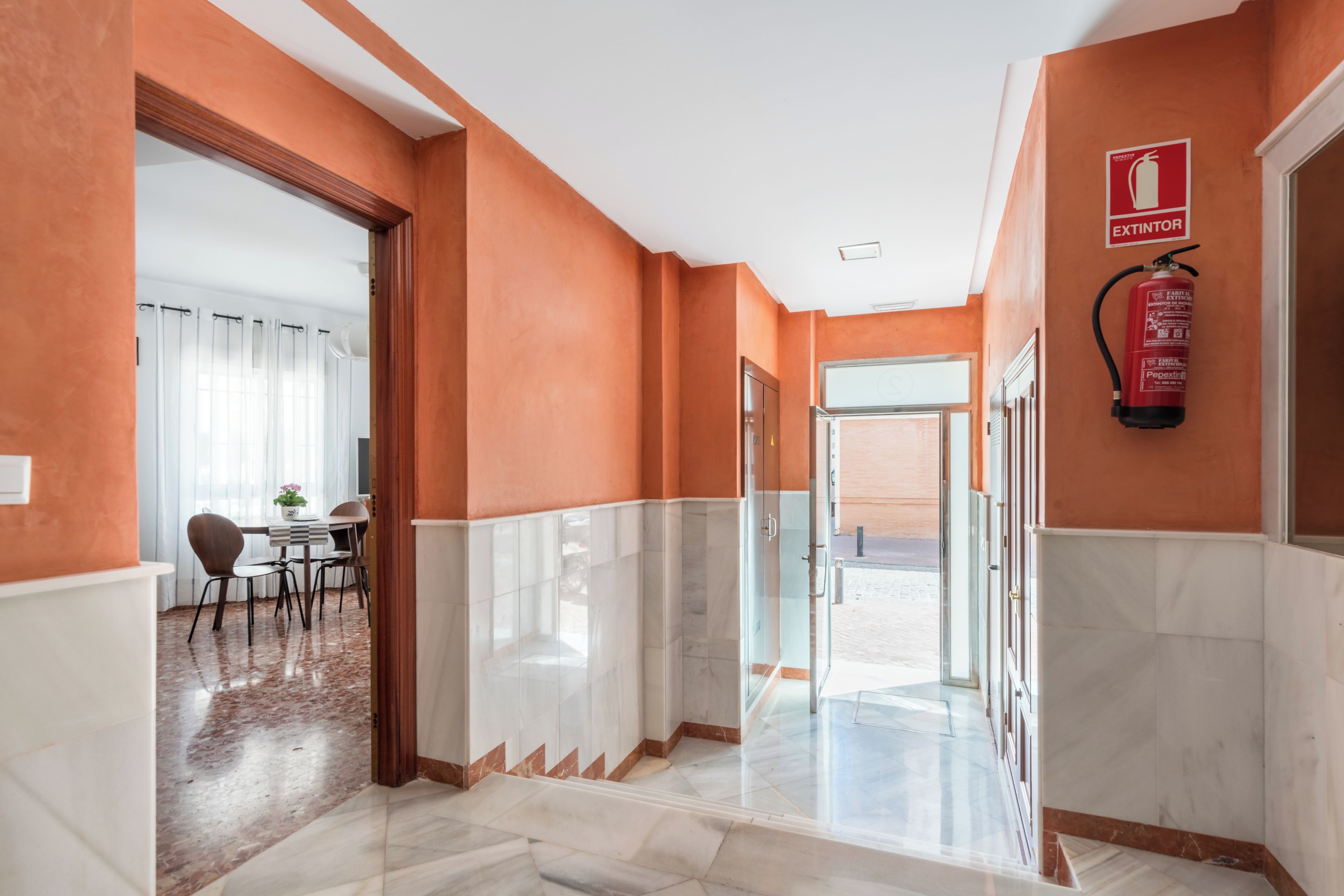 A hotel corridor with a fire extinguisher mounted on the wall, a glass door leading to a room, and a dining area with a table and chairs.