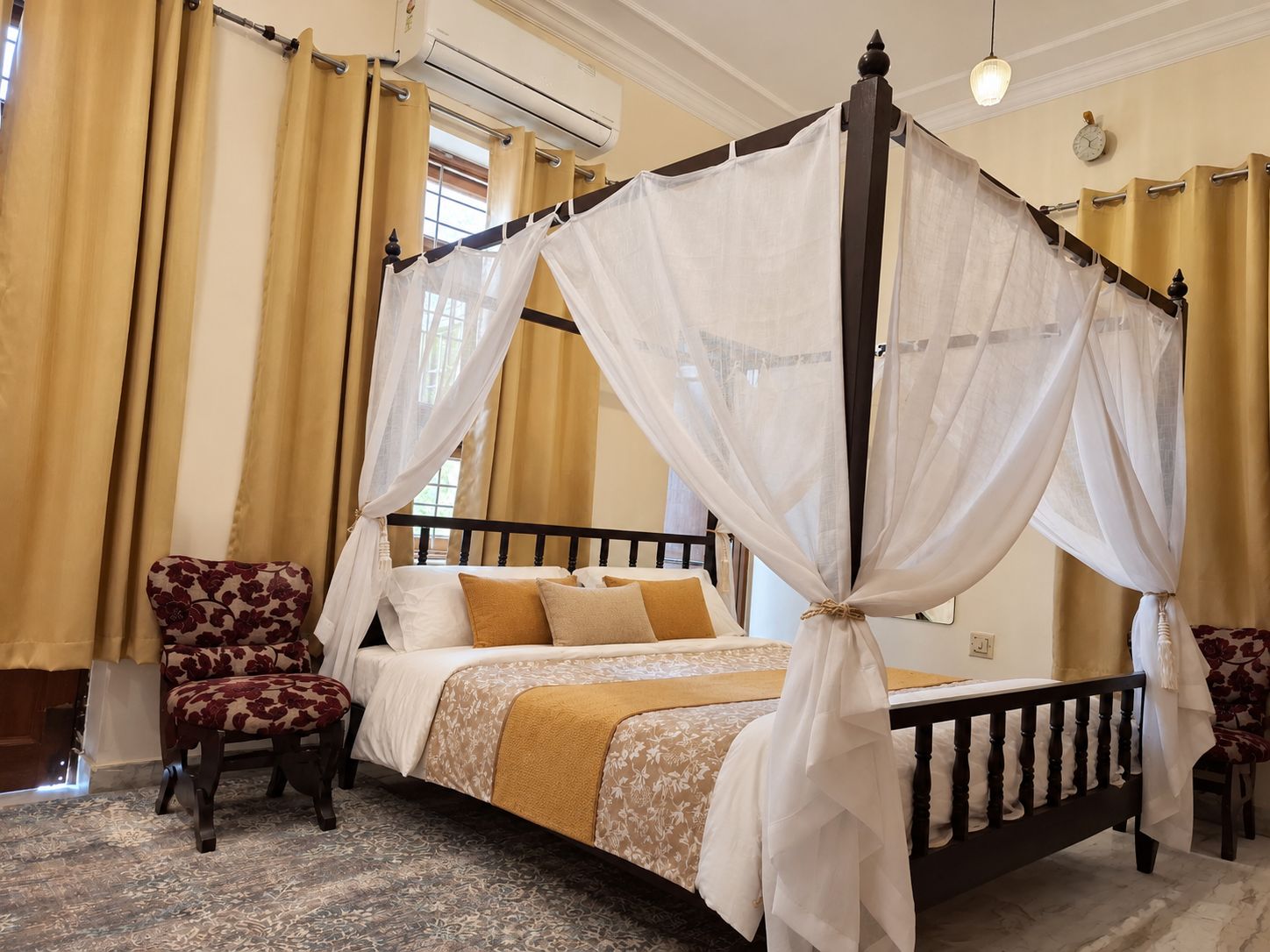 A four-poster canopy bed with a floral-patterned chair beside it, a second chair, and a carpeted floor.