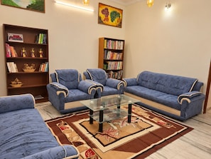 Een woonkamer met een blauwe bank, twee fauteuils, een glazen salontafel en een vloerkleed met een patroon. Er staan twee boekenplanken vol boeken en trofeeën.