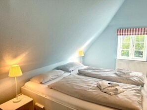 Une chambre avec deux lits, une fenêtre ornée d’une lambrequin à carreaux et une lampe placée sur le côté gauche.