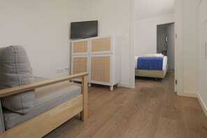 Een moderne, minimalistische kamer met een grijze bank, een klein houten tafeltje, een bed met een blauwe deken en een televisie bevestigd aan een kast.
