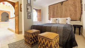 Een slaapkamer met een groot bed, twee geweven krukken, een gestructureerd hoofdeinde en uitzicht op een binnenplaats.