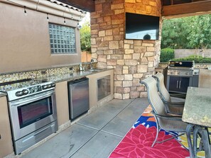 An outdoor kitchen with a stone wall, a built-in oven, a grill, and a dining area with a colorful rug. - AllStar Retreat! PickleBall/Soccer/Pool/Waterslide/Spa/Splash Pad/Putting Greens (Queen Creek)