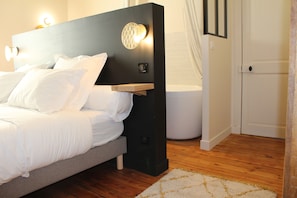 A modern bedroom with a bed, bedside lamps, a nightstand, and a bathroom with a bathtub. - Maison Caju (La Rochelle)