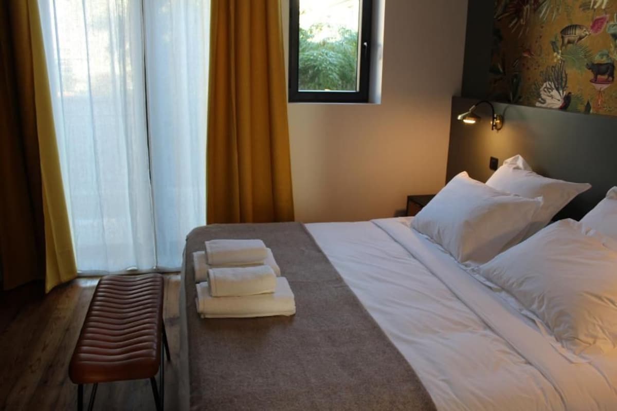 A hotel room with a bed, a bench, a window with curtains, and a wall-mounted lamp.