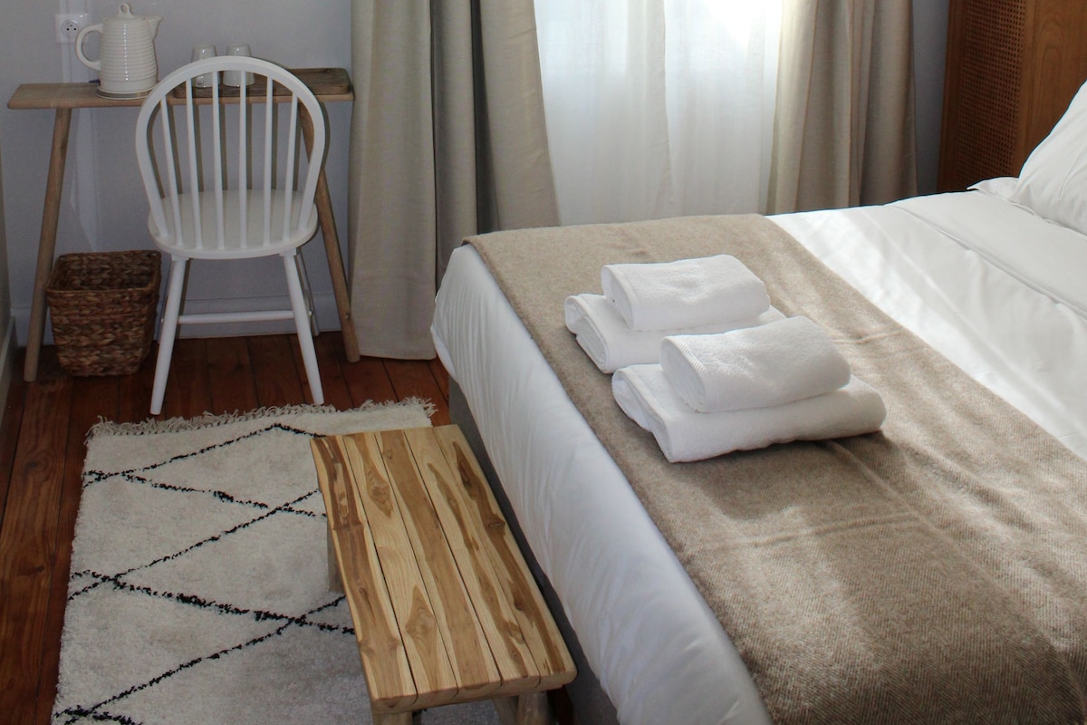 A hotel room with a bed, a wooden bench, a white chair, a television, and a window with curtains.