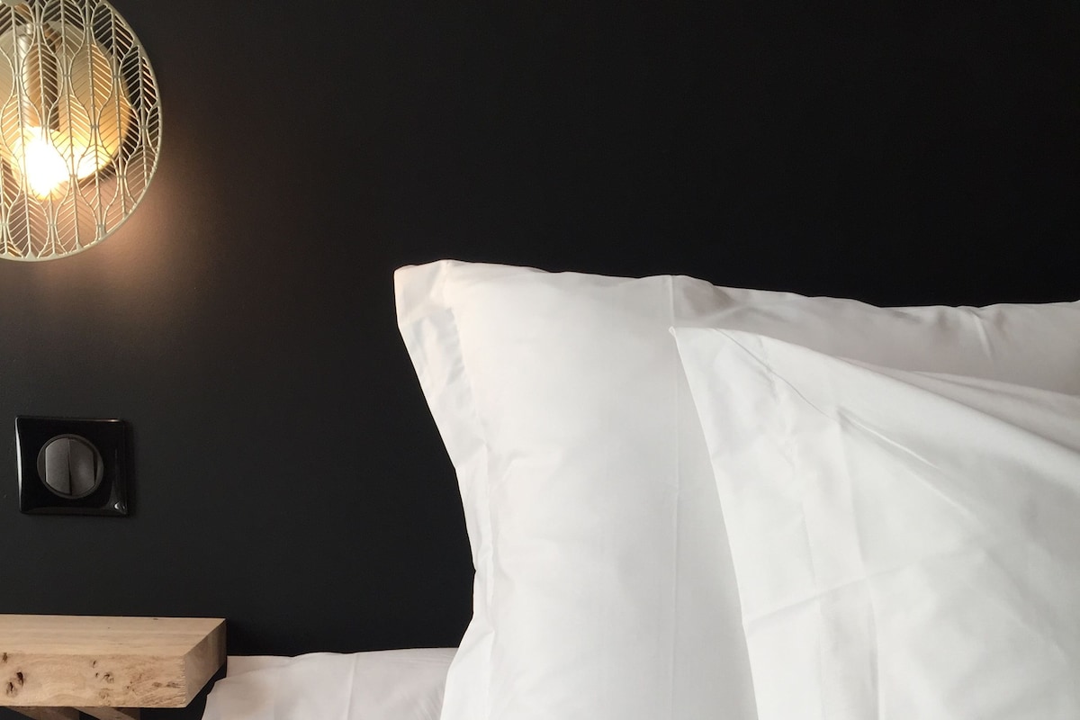 A bed with white bedding, a wooden headboard, and a wall-mounted light fixture.