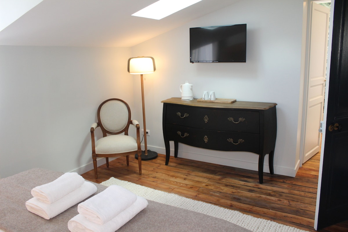 A room with a wooden floor, a white chair, a bedside table with a lamp, a TV mounted on the wall, and a door.