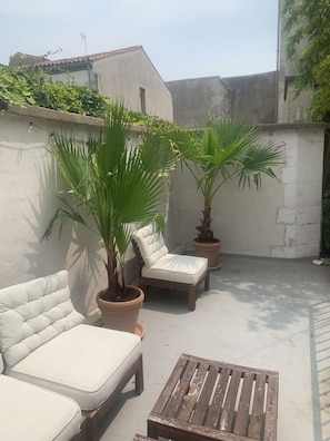 A patio with two white cushioned chairs, a wooden table, and two potted palm trees.