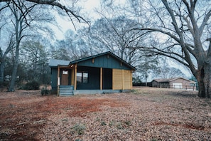 Eine kleine, eingeschossige Holzhütte mit Veranda und Überdachung, umgeben von Bäumen und einem mit Laub bedeckten Boden.