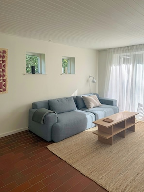 A modern living room with a sectional sofa, a wooden coffee table, and a small window with a view of trees.