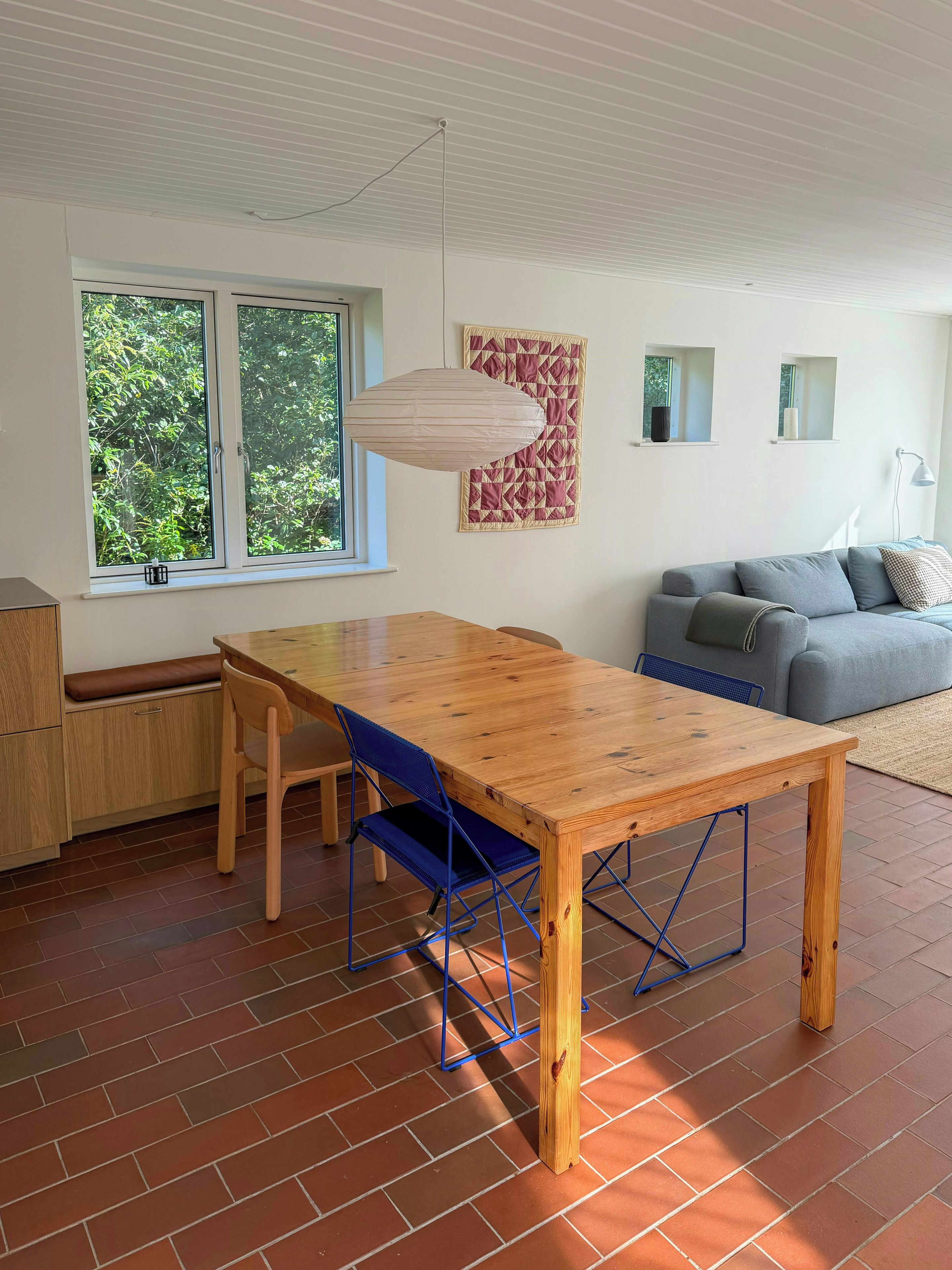 A dining area with a wooden table and blue chairs, a sofa, and a quilt on the wall.