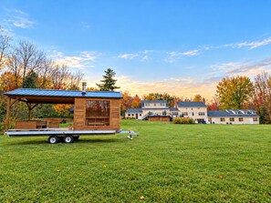 A mobile home with a metal roof and wooden walls, parked on a grassy field with a house in the background. - The Wiley | Mountain Retreat with Hot Tub & Sauna (Peru)