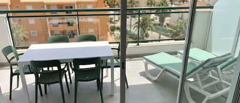 A balcony with a table and chairs, a lounge chair, and a view of the sea and buildings.