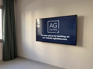 A flat-screen TV mounted on a wall displaying a logo and text for AG Suites, advertising the best prices by booking on their website. - The Emerald Penthouses (Portimão)