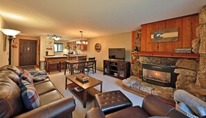 Una sala de estar con chimenea de piedra, televisión y una cocina al fondo.