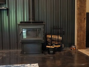 Una estufa de leña con puerta de cristal, un cubo negro con asa y una caja de iniciadores de fuego sobre el suelo.