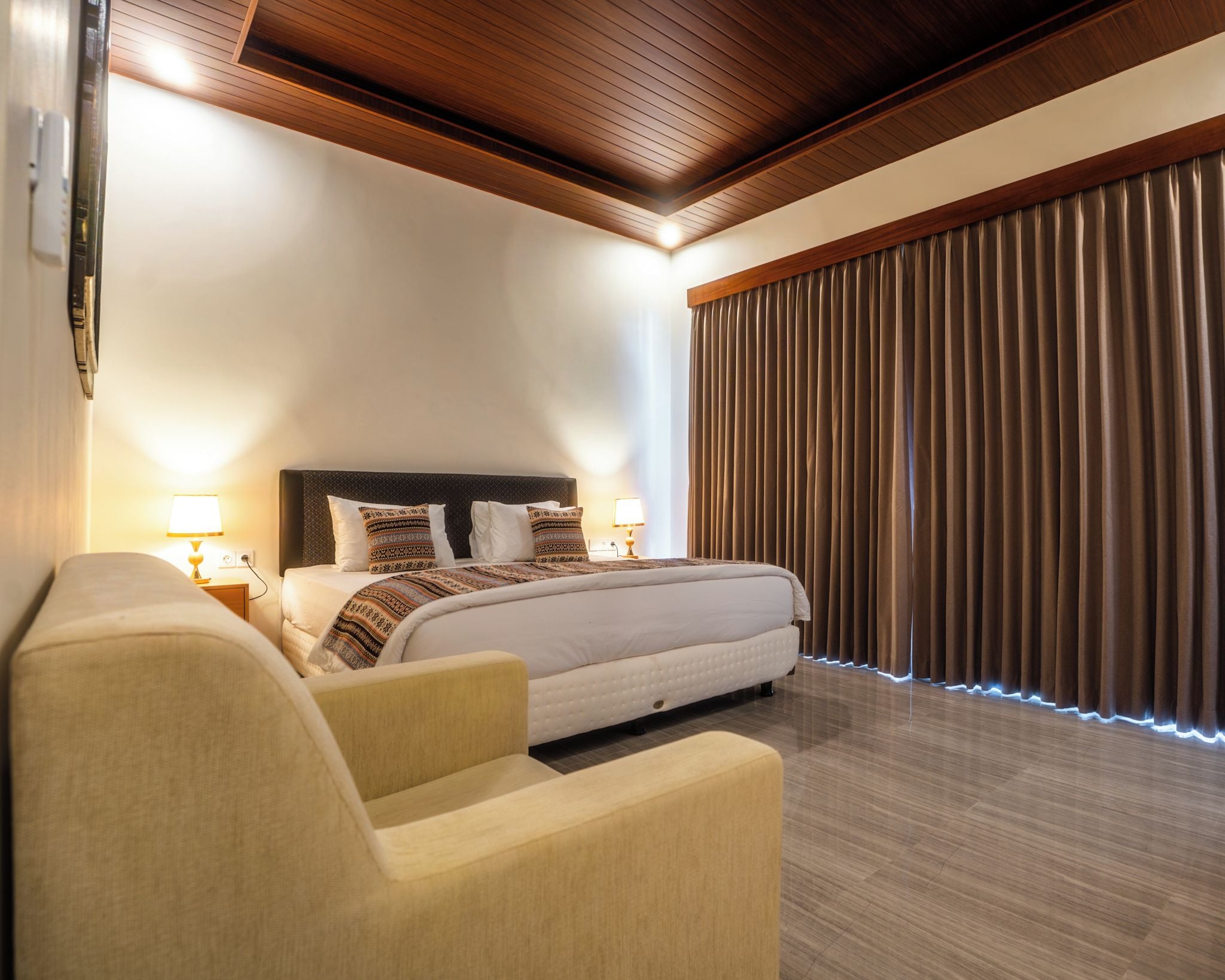 A hotel room with a bed, bedside lamps, a sofa, a wooden ceiling, and a large window with curtains.