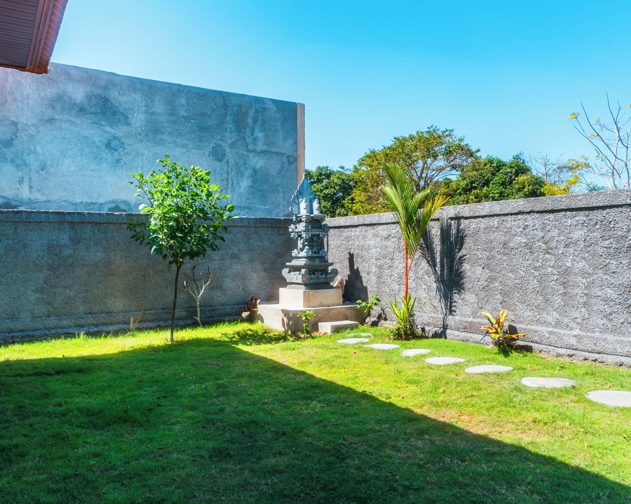 Pátio com muro de pedra, uma pequena árvore, uma estátua e área gramada com pedras para caminhar.