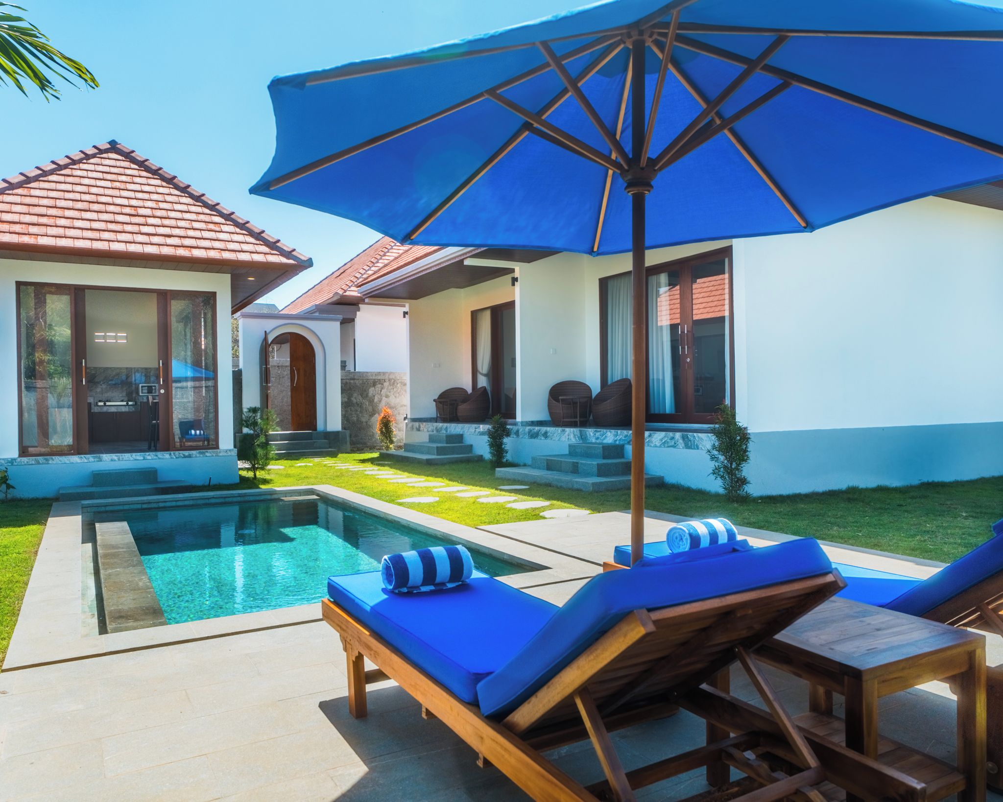 A poolside area with lounge chairs and a large umbrella, a house with a tiled roof, and a well-maintained lawn.